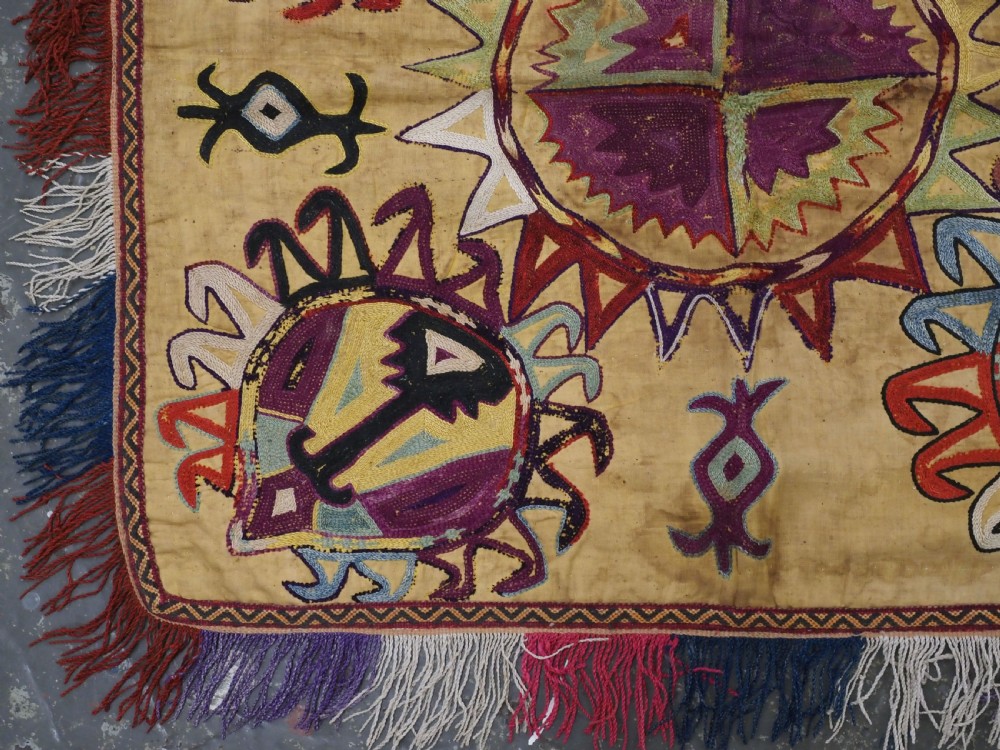 A square antique Uzbek Lakai embroidered textile featuring central circular and solar motifs in purple, red, and turquoise on a yellow fabric ground, finished with a multi-coloured fringed border. View IV