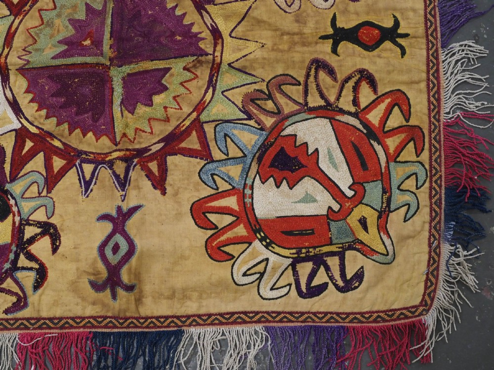 A square antique Uzbek Lakai embroidered textile featuring central circular and solar motifs in purple, red, and turquoise on a yellow fabric ground, finished with a multi-coloured fringed border. View V