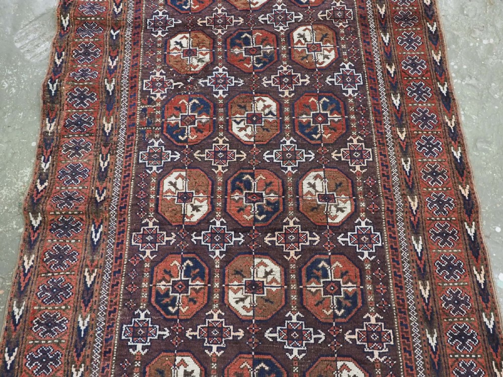 An antique woven wool Baluch rug featuring repeating octagonal Turkmen gul motifs in dark brown, terracotta, navy blue, and ivory with geometric borders. View III
