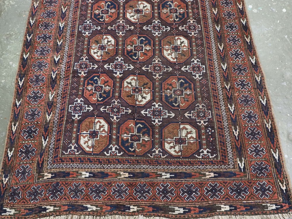 An antique woven wool Baluch rug featuring repeating octagonal Turkmen gul motifs in dark brown, terracotta, navy blue, and ivory with geometric borders. View IV