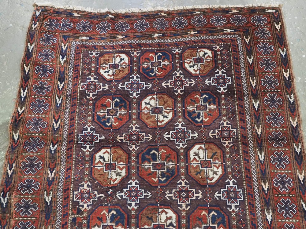An antique woven wool Baluch rug featuring repeating octagonal Turkmen gul motifs in dark brown, terracotta, navy blue, and ivory with geometric borders. View V