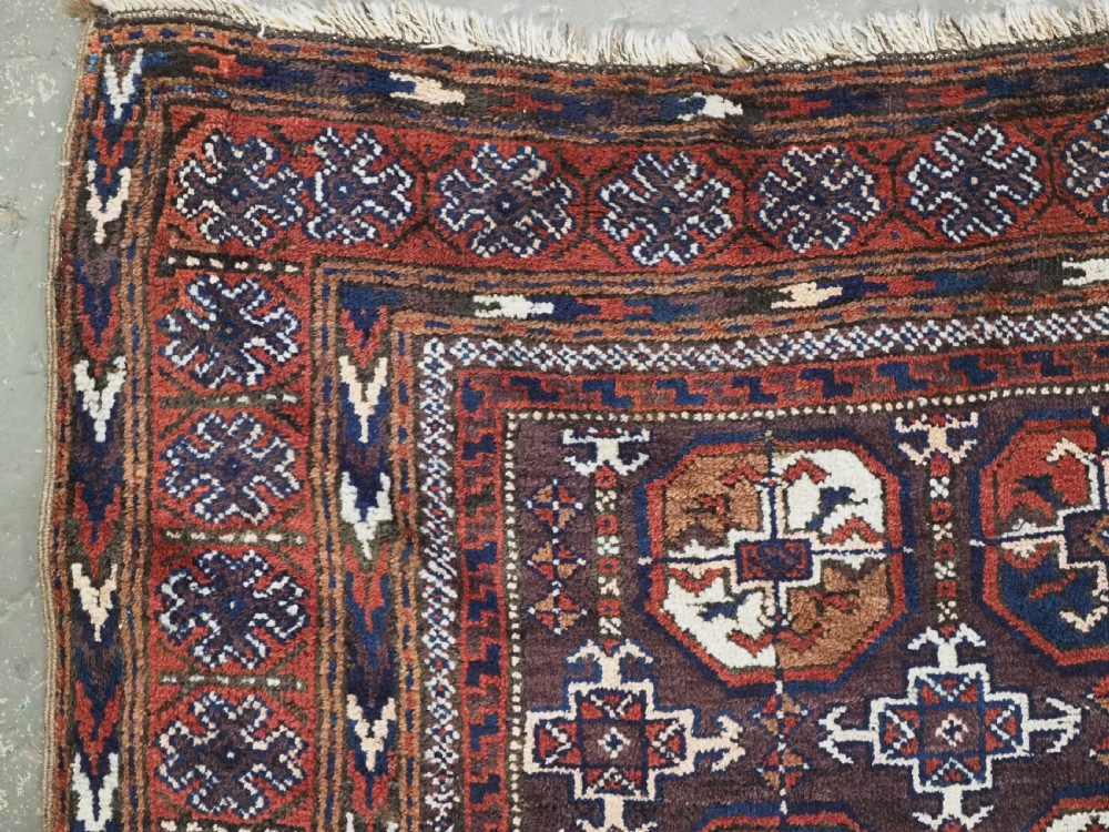 An antique woven wool Baluch rug featuring repeating octagonal Turkmen gul motifs in dark brown, terracotta, navy blue, and ivory with geometric borders. View VI