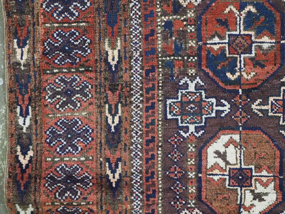 An antique woven wool Baluch rug featuring repeating octagonal Turkmen gul motifs in dark brown, terracotta, navy blue, and ivory with geometric borders. View VII