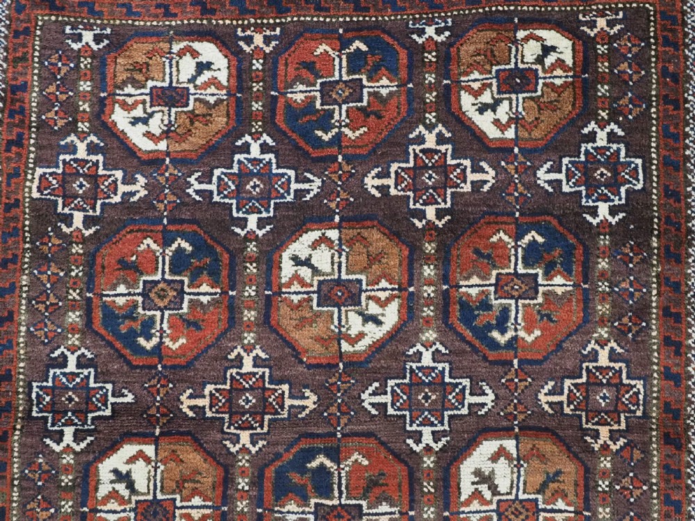 An antique woven wool Baluch rug featuring repeating octagonal Turkmen gul motifs in dark brown, terracotta, navy blue, and ivory with geometric borders. View VIII