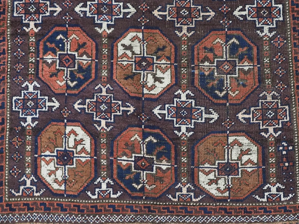 An antique woven wool Baluch rug featuring repeating octagonal Turkmen gul motifs in dark brown, terracotta, navy blue, and ivory with geometric borders. View IX