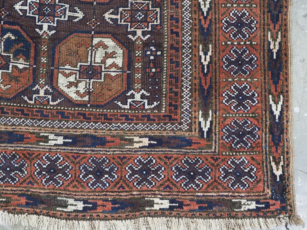 An antique woven wool Baluch rug featuring repeating octagonal Turkmen gul motifs in dark brown, terracotta, navy blue, and ivory with geometric borders. View X