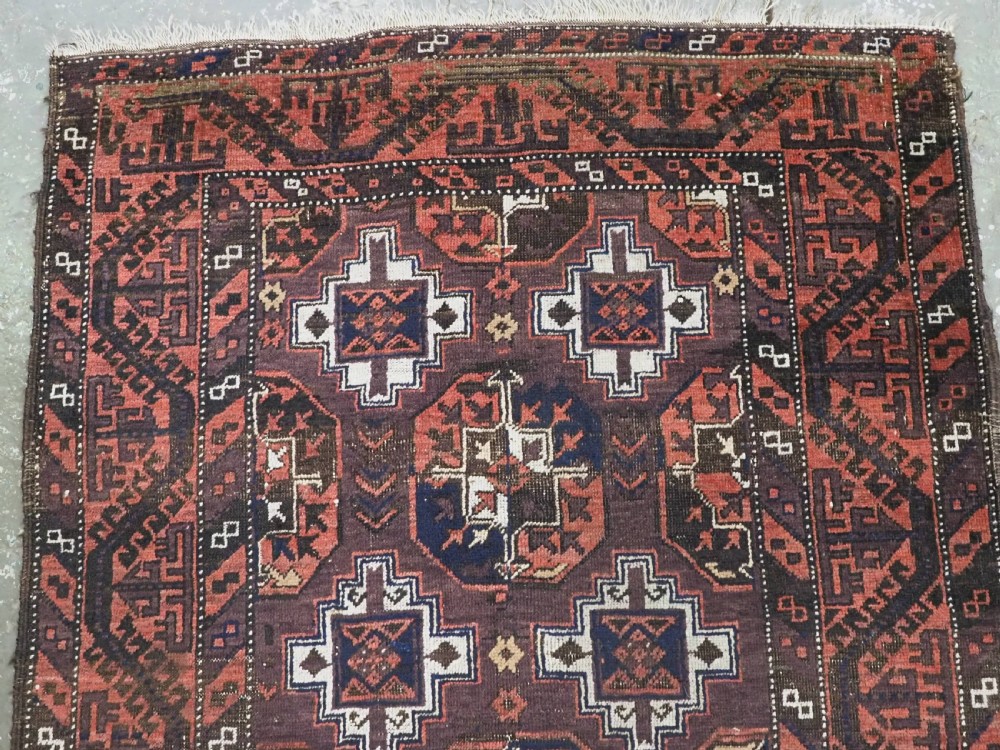 An antique hand-knotted Baluch wool rug displaying repeating geometric cruciform and octagonal motifs in terracotta red, navy blue, and dark brown tones. View III