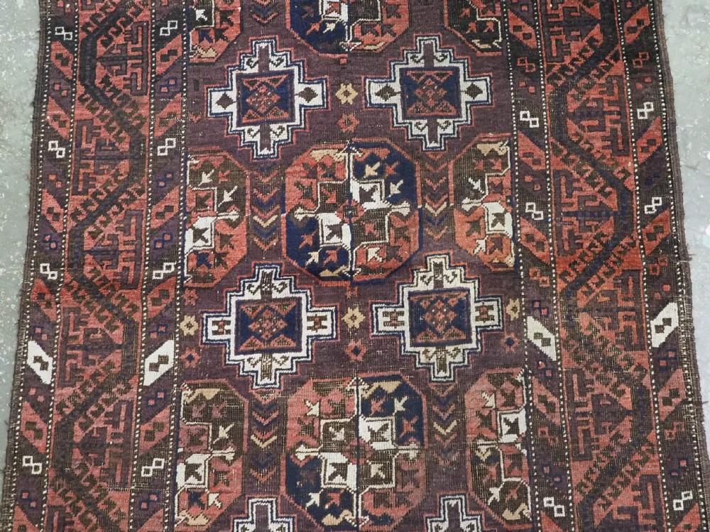 An antique hand-knotted Baluch wool rug displaying repeating geometric cruciform and octagonal motifs in terracotta red, navy blue, and dark brown tones. View IV