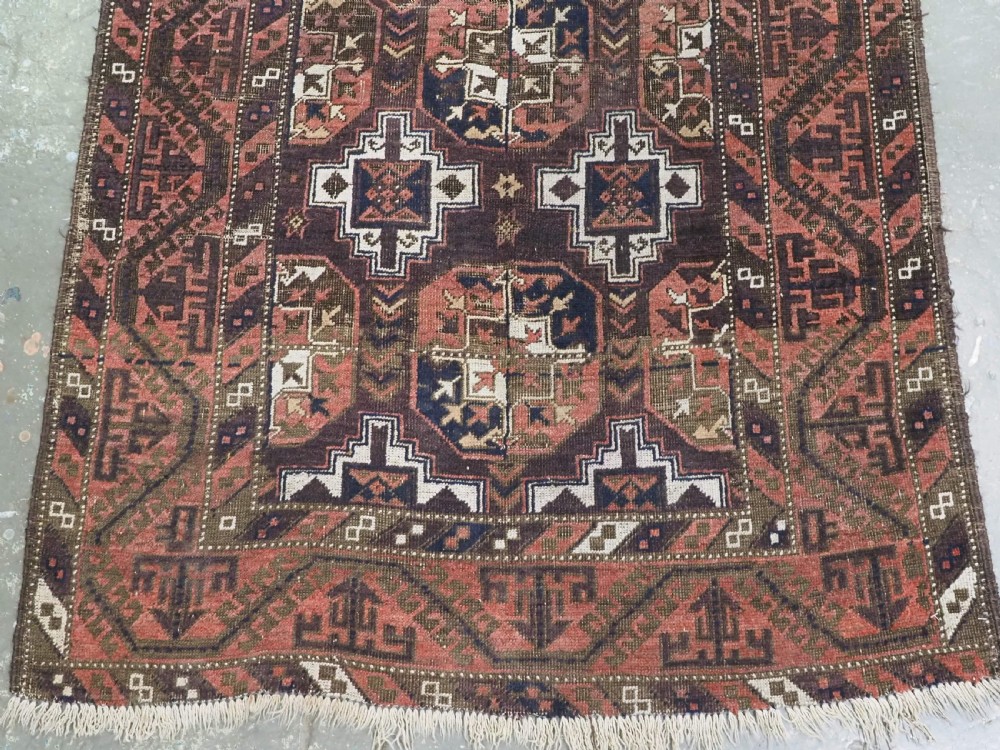 An antique hand-knotted Baluch wool rug displaying repeating geometric cruciform and octagonal motifs in terracotta red, navy blue, and dark brown tones. View V