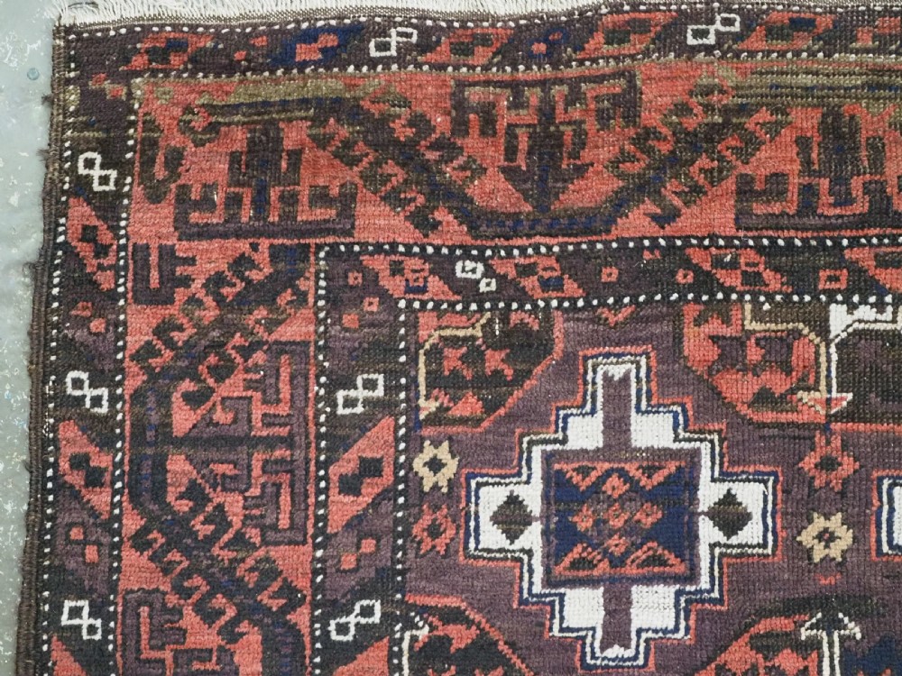 An antique hand-knotted Baluch wool rug displaying repeating geometric cruciform and octagonal motifs in terracotta red, navy blue, and dark brown tones. View VI