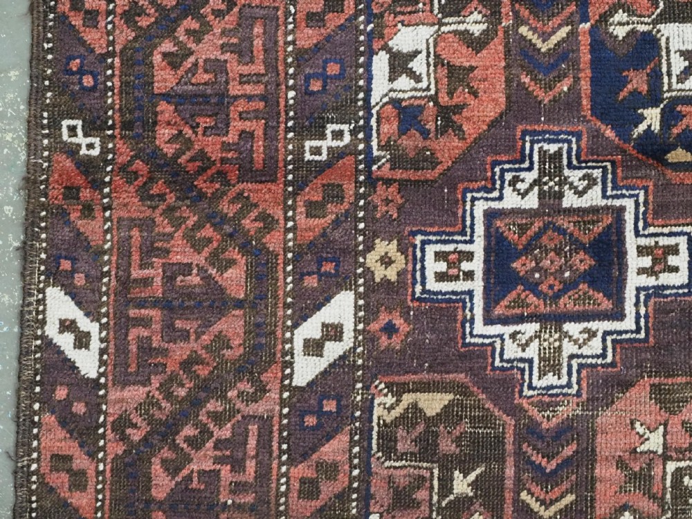 An antique hand-knotted Baluch wool rug displaying repeating geometric cruciform and octagonal motifs in terracotta red, navy blue, and dark brown tones. View VII