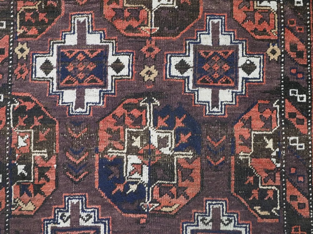 An antique hand-knotted Baluch wool rug displaying repeating geometric cruciform and octagonal motifs in terracotta red, navy blue, and dark brown tones. View VIII