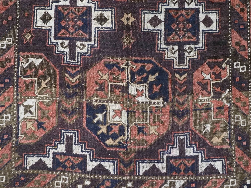An antique hand-knotted Baluch wool rug displaying repeating geometric cruciform and octagonal motifs in terracotta red, navy blue, and dark brown tones. View IX