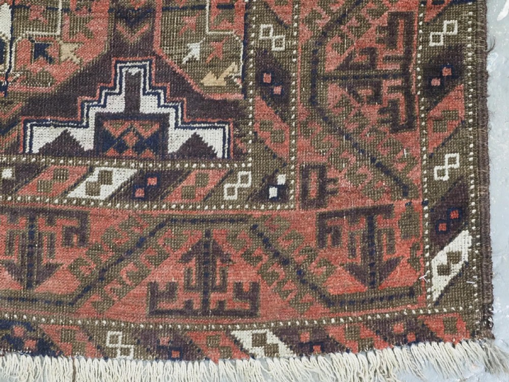 An antique hand-knotted Baluch wool rug displaying repeating geometric cruciform and octagonal motifs in terracotta red, navy blue, and dark brown tones. View X