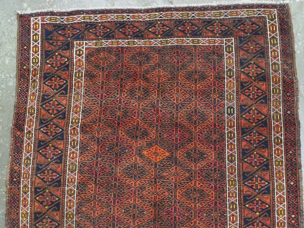 An antique rectangular Baluch wool rug featuring a terracotta geometric lattice pattern on a dark field surrounded by framed patterned borders. View III