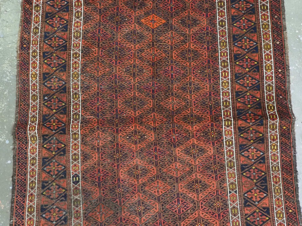 An antique rectangular Baluch wool rug featuring a terracotta geometric lattice pattern on a dark field surrounded by framed patterned borders. View IV