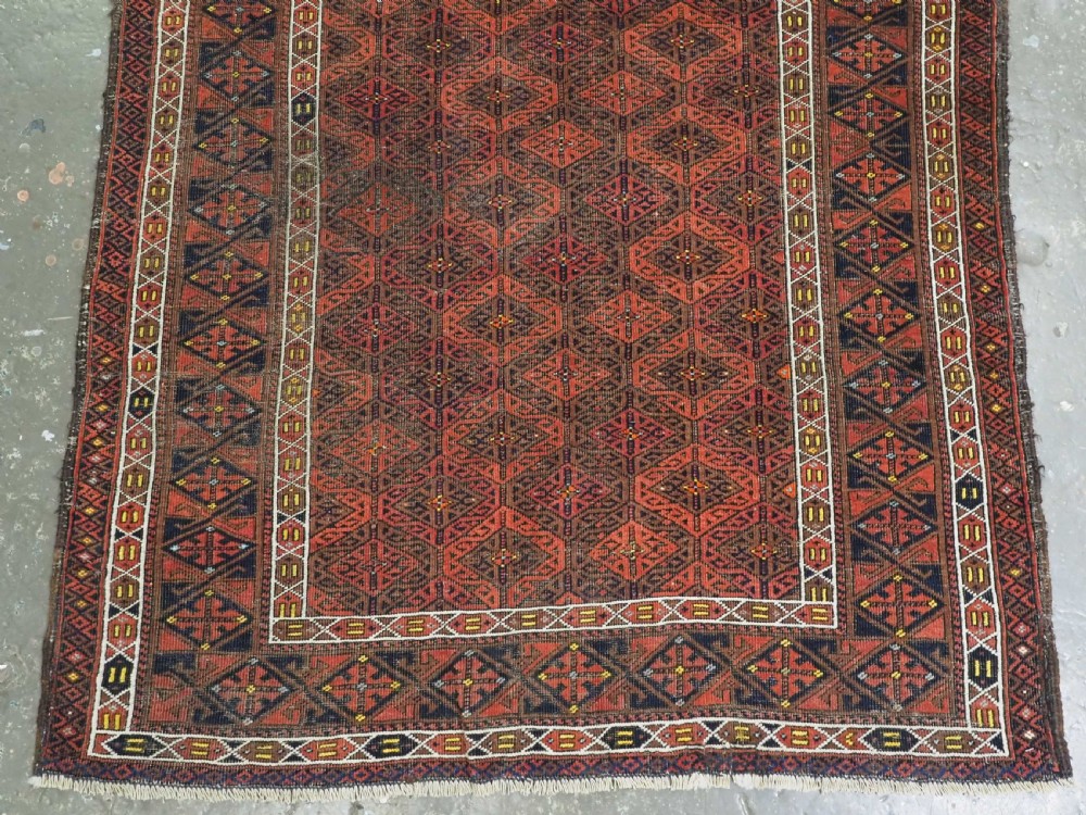 An antique rectangular Baluch wool rug featuring a terracotta geometric lattice pattern on a dark field surrounded by framed patterned borders. View V
