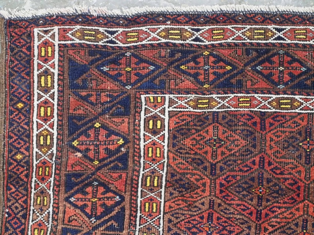 An antique rectangular Baluch wool rug featuring a terracotta geometric lattice pattern on a dark field surrounded by framed patterned borders. View VI