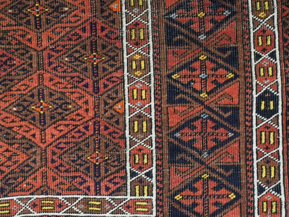 An antique rectangular Baluch wool rug featuring a terracotta geometric lattice pattern on a dark field surrounded by framed patterned borders. View XII