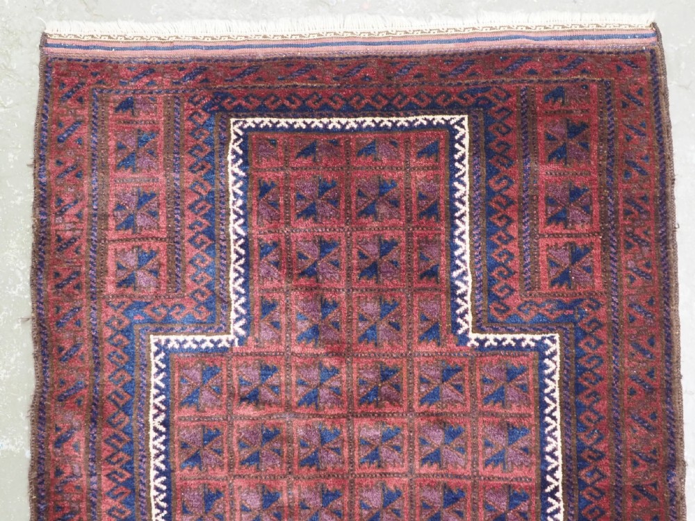 An antique hand-knotted Baluch wool prayer rug featuring a central mihrab arch motif with repeat geometric lotus leaf patterns in deep red, navy blue, and white. View III