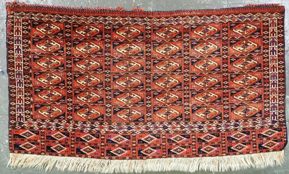 A hand-knotted wool Tekke Turkmen chuval face with repeating geometric aina gul motifs in red, navy blue, and ivory tones, finished with a fringed lower border.