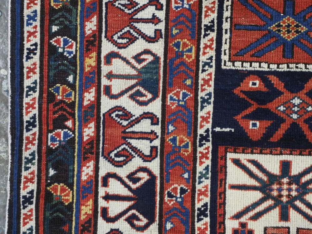 An antique hand-knotted Caucasian Shirvan wool rug featuring geometric stepped medallions on a deep blue field with red and cream patterned borders. View VII