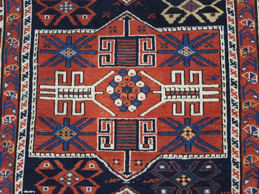 An antique hand-knotted Caucasian Shirvan wool rug featuring geometric stepped medallions on a deep blue field with red and cream patterned borders. View IX