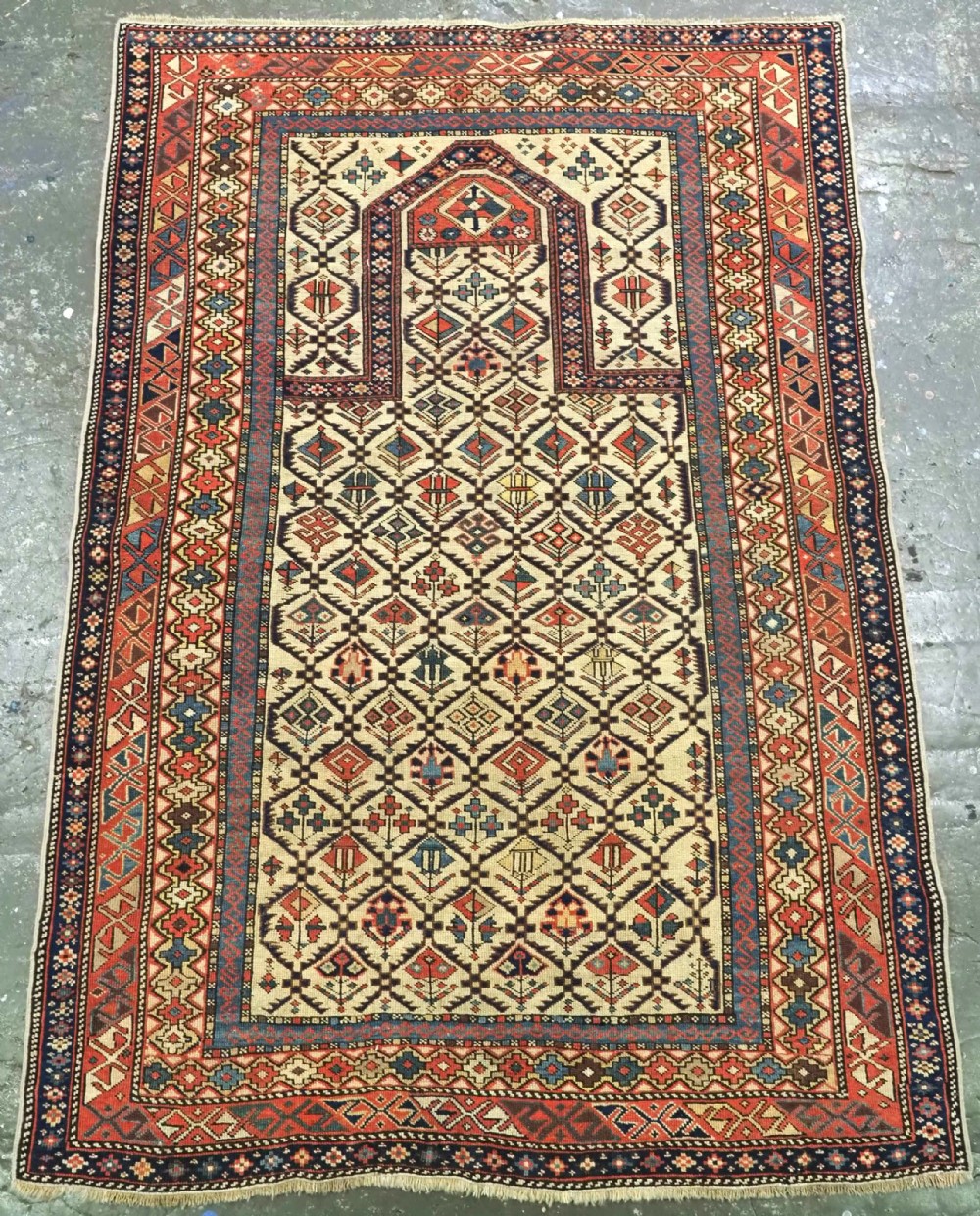An antique Caucasian Shirvan prayer rug featuring an ivory ground decorated with a geometric floral lattice pattern and a arched mihrab.