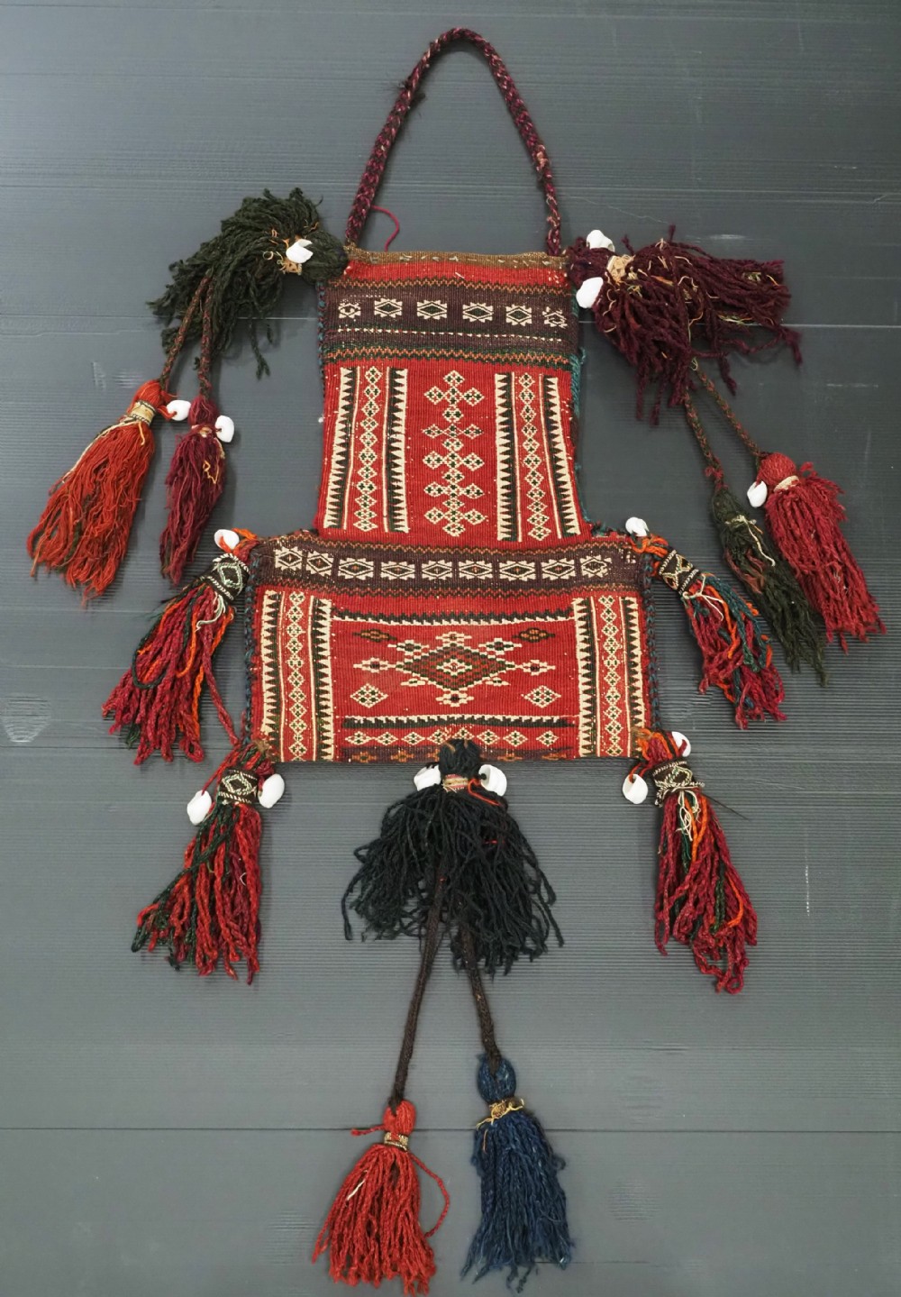 An antique woven wool salt bag featuring intricate red and dark brown geometric patterns, decorated with braided wool tassels and white cowrie shells.