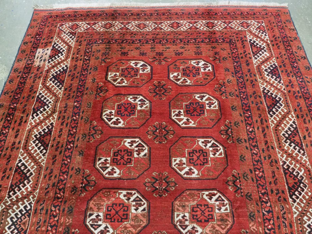 A rectangular hand-knotted wool carpet featuring a deep red field decorated with two columns of octagonal gul motifs surrounded by geometric borders. View III