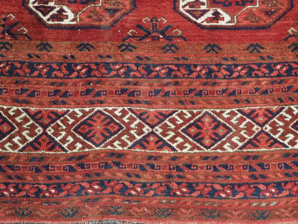 A rectangular hand-knotted wool carpet featuring a deep red field decorated with two columns of octagonal gul motifs surrounded by geometric borders. View X