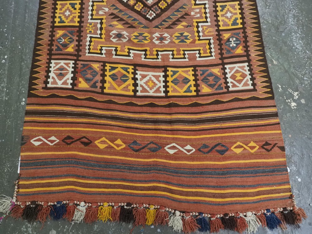 A long flat-woven Afghan kilim runner carpet featuring geometric diamond motifs in terracotta, brown, yellow, and ivory shades with a tasseled fringe. View VI