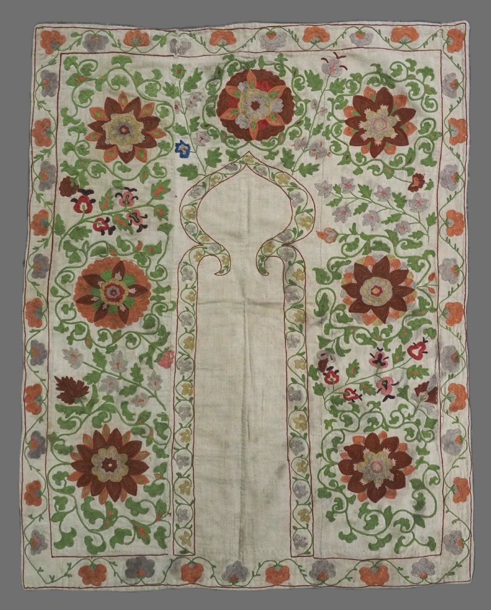 A cream woven cotton textile panel decorated with intricate silk embroidery depicting a pointed arch niche surrounded by floral rosettes and vine tendrils in terracotta and green tones.