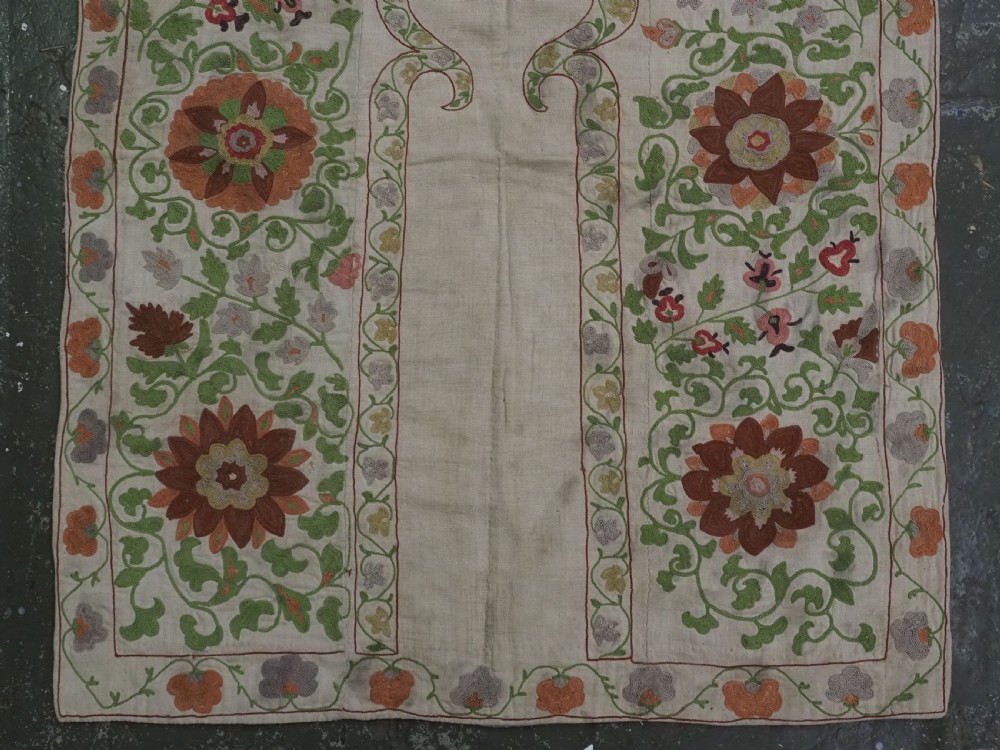 A cream woven cotton textile panel decorated with intricate silk embroidery depicting a pointed arch niche surrounded by floral rosettes and vine tendrils in terracotta and green tones. View IV