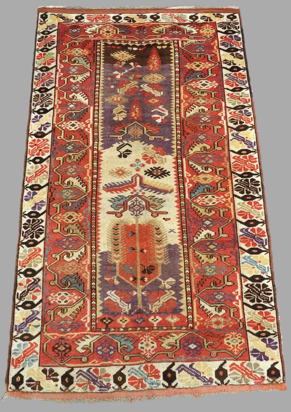 An antique hand-knotted Turkish Milas wool prayer rug featuring a central ivory prayer niche flanked by geometric borders in terracotta, purple, and cream tones.