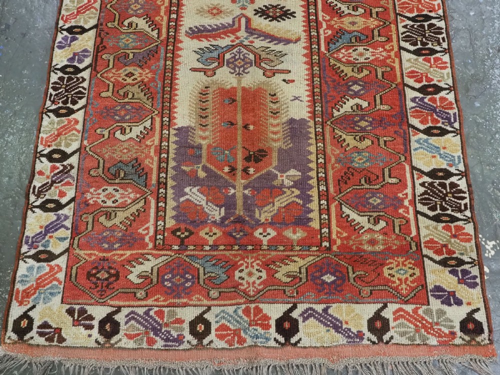 An antique hand-knotted Turkish Milas wool prayer rug featuring a central ivory prayer niche flanked by geometric borders in terracotta, purple, and cream tones. View IV