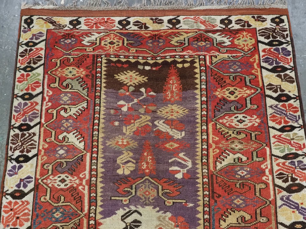 An antique hand-knotted Turkish Milas wool prayer rug featuring a central ivory prayer niche flanked by geometric borders in terracotta, purple, and cream tones. View V