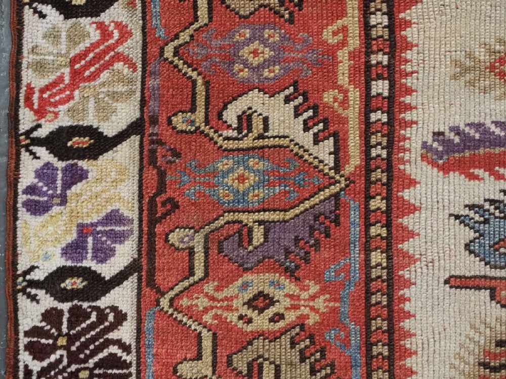 An antique hand-knotted Turkish Milas wool prayer rug featuring a central ivory prayer niche flanked by geometric borders in terracotta, purple, and cream tones. View VI