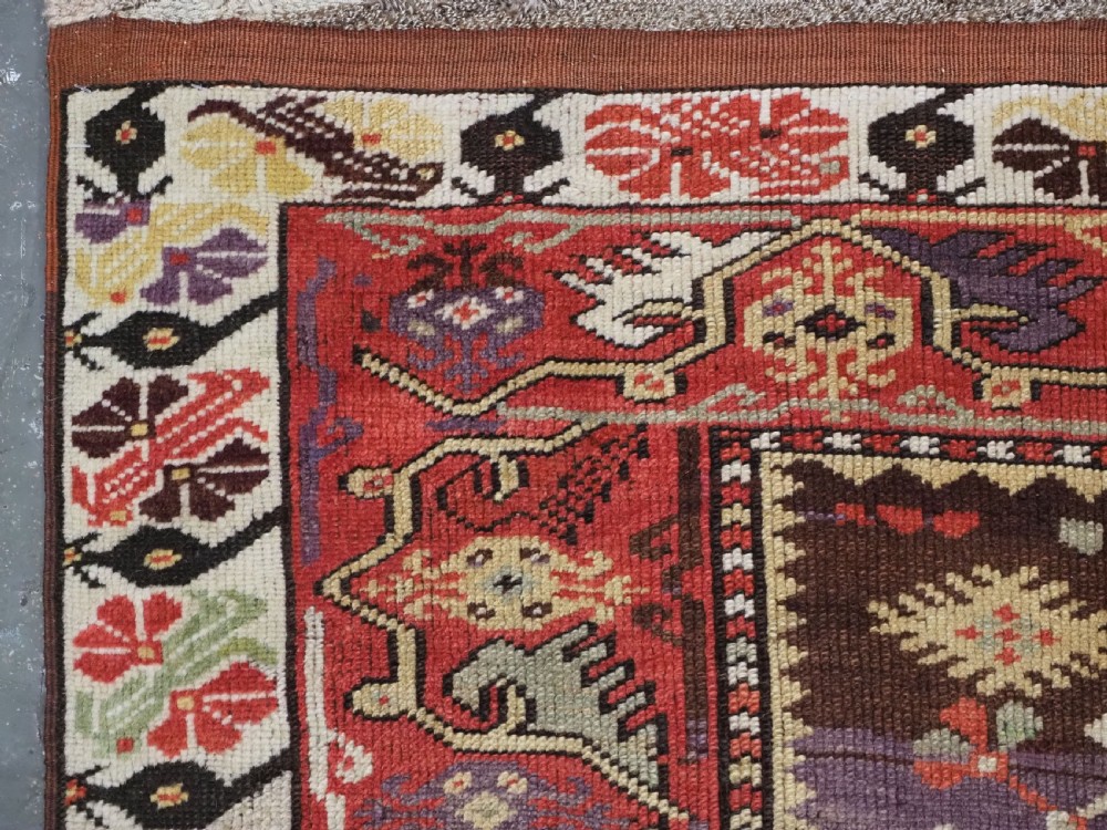 An antique hand-knotted Turkish Milas wool prayer rug featuring a central ivory prayer niche flanked by geometric borders in terracotta, purple, and cream tones. View VII