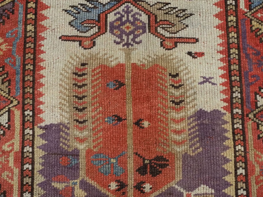 An antique hand-knotted Turkish Milas wool prayer rug featuring a central ivory prayer niche flanked by geometric borders in terracotta, purple, and cream tones. View X