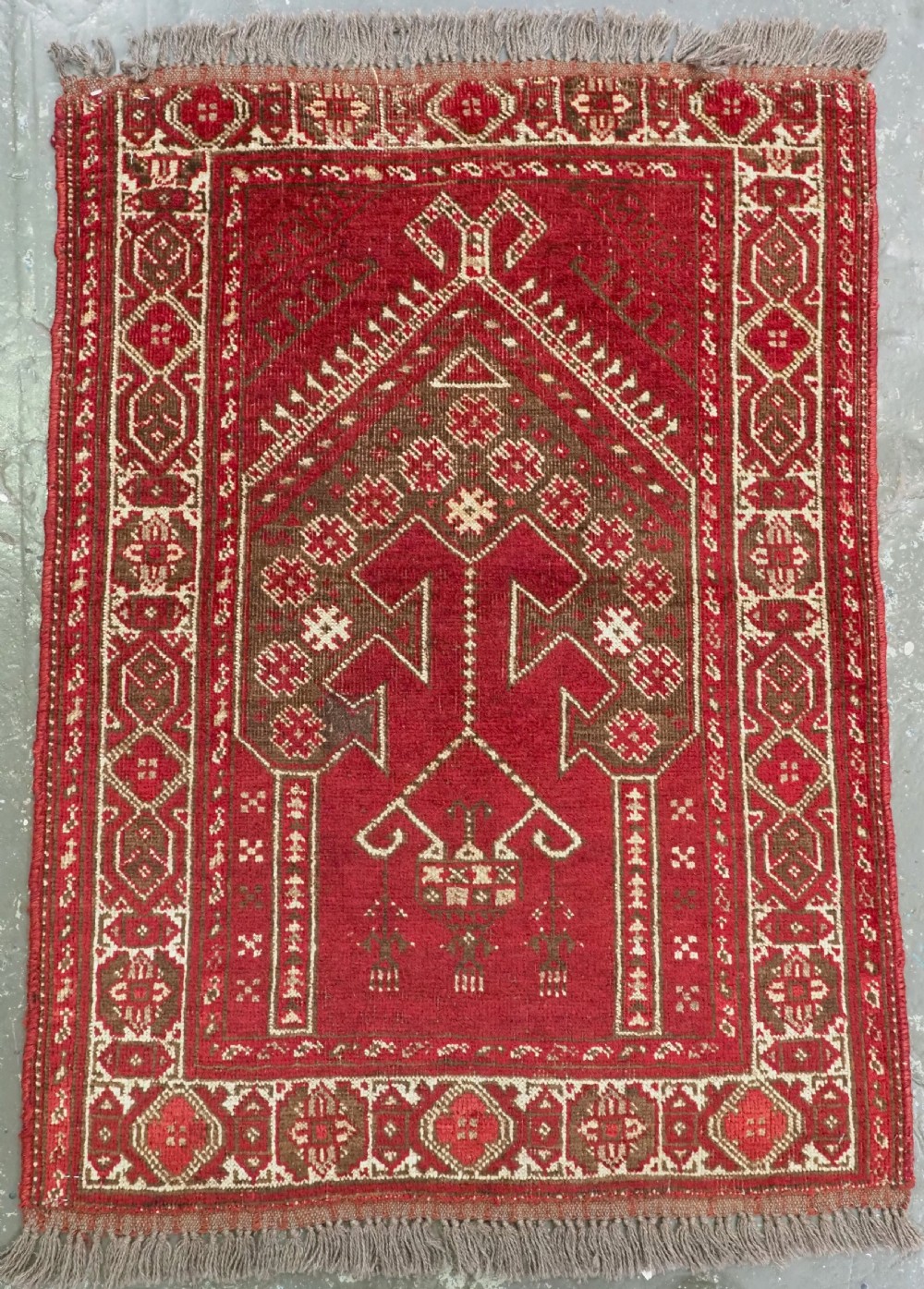 An antique hand-knotted wool Afghan prayer rug featuring a deep red field, a prominent central arch motif, and geometric patterned borders in brown and cream.