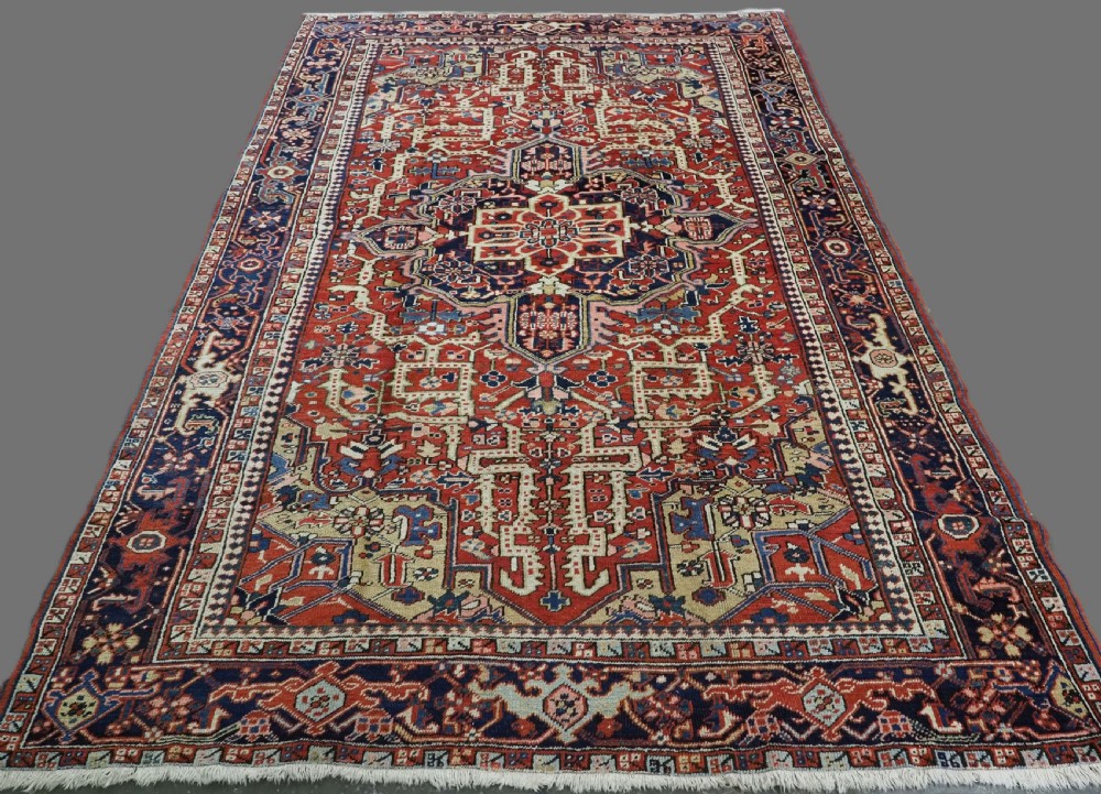 An antique Persian Heriz wool carpet featuring a central geometric medallion, stylized floral motifs in red, blue, and cream, framed by a complex multi-tiered border.