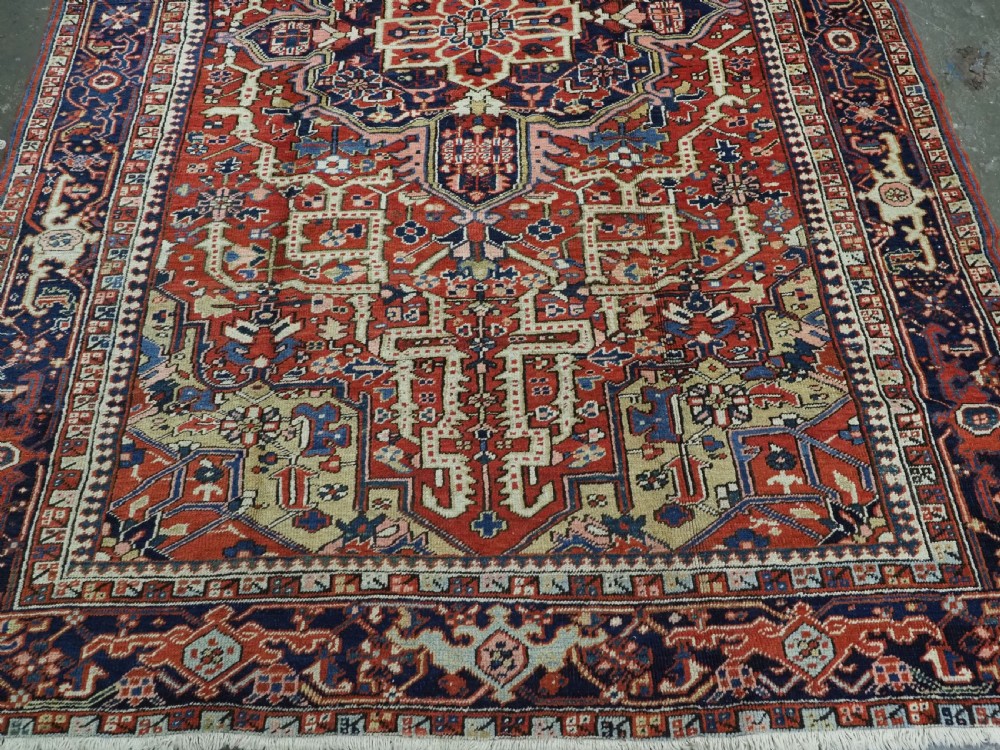 An antique Persian Heriz wool carpet featuring a central geometric medallion, stylized floral motifs in red, blue, and cream, framed by a complex multi-tiered border. View V