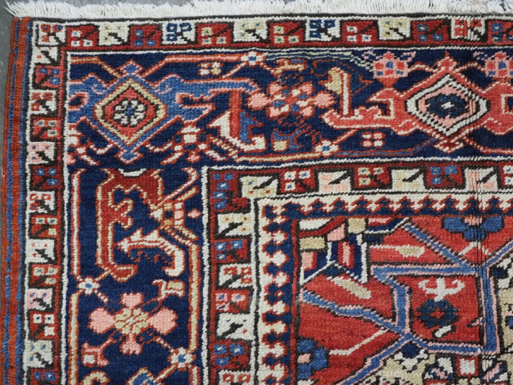 An antique Persian Heriz wool carpet featuring a central geometric medallion, stylized floral motifs in red, blue, and cream, framed by a complex multi-tiered border. View VI