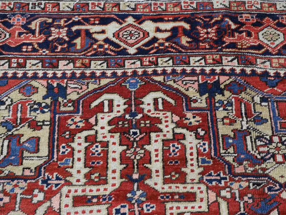 An antique Persian Heriz wool carpet featuring a central geometric medallion, stylized floral motifs in red, blue, and cream, framed by a complex multi-tiered border. View IX