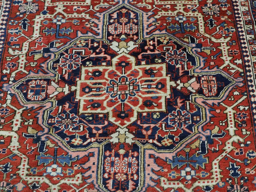 An antique Persian Heriz wool carpet featuring a central geometric medallion, stylized floral motifs in red, blue, and cream, framed by a complex multi-tiered border. View X
