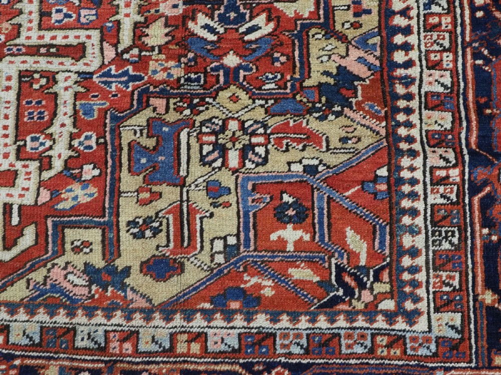 An antique Persian Heriz wool carpet featuring a central geometric medallion, stylized floral motifs in red, blue, and cream, framed by a complex multi-tiered border. View XII