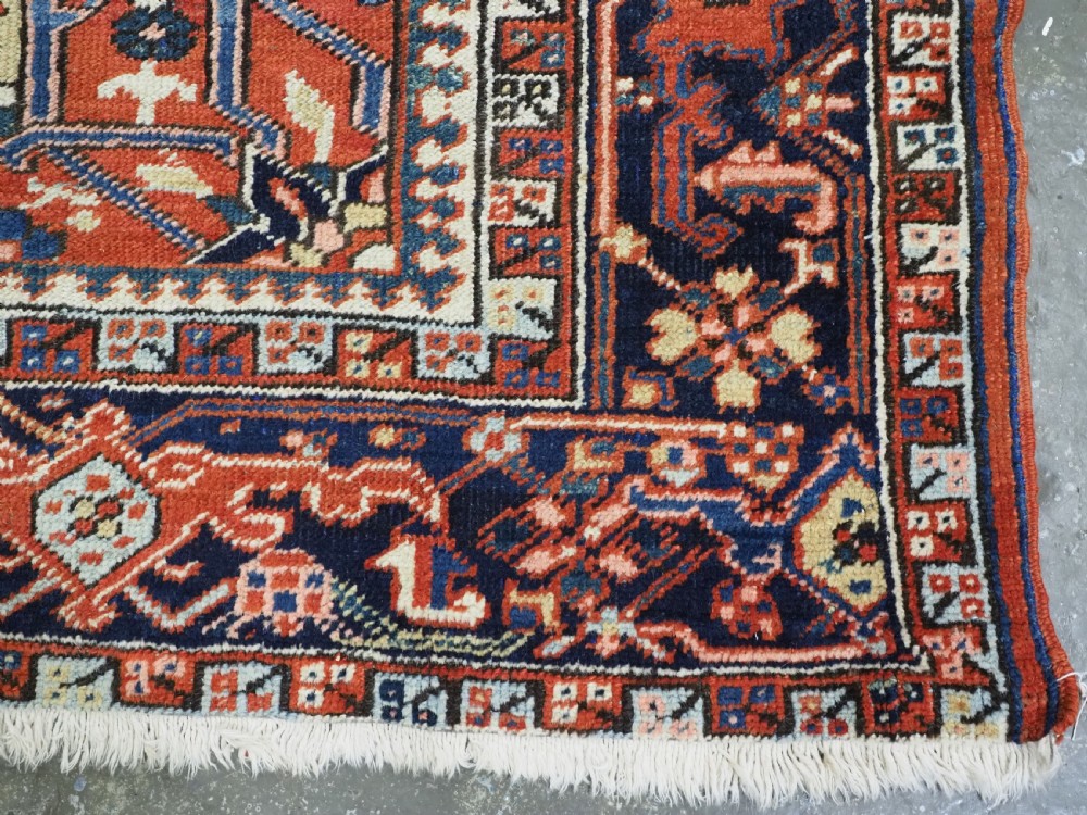 An antique Persian Heriz wool carpet featuring a central geometric medallion, stylized floral motifs in red, blue, and cream, framed by a complex multi-tiered border. View XIII