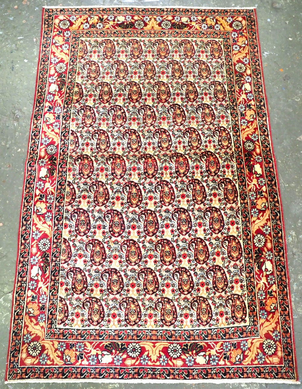An antique Kirman rug featuring a repeat floral boteh pattern on a cream field enclosed by a detailed red and cream floral border.
