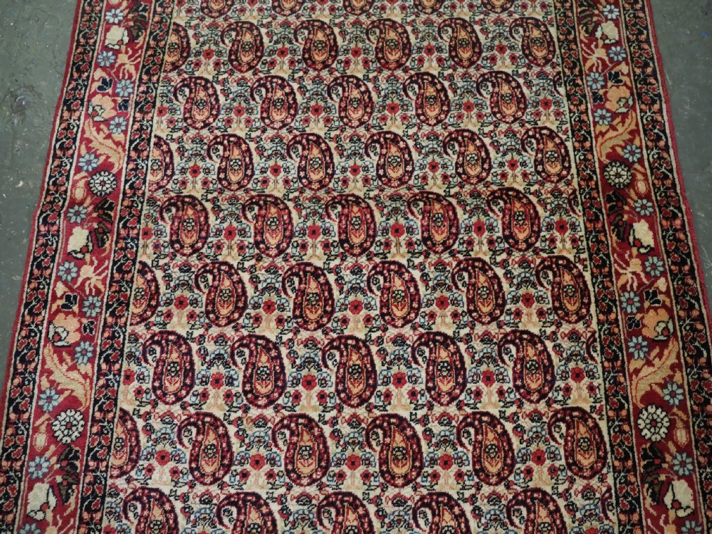 An antique Kirman rug featuring a repeat floral boteh pattern on a cream field enclosed by a detailed red and cream floral border. View IV
